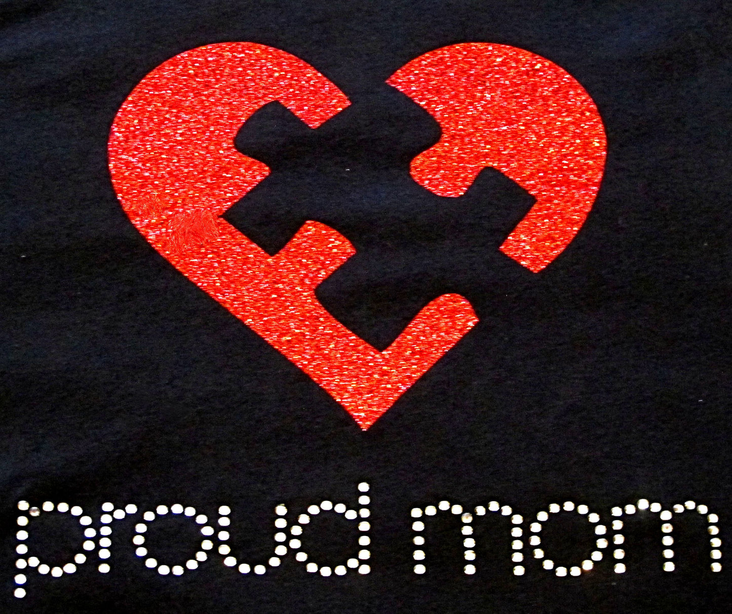 Embrace Difference Tees -Proud Mom - Tumble into Love close up of tee shirt image.  A red glitter heart with a black puzzle piece in the center.  Proud mom is spelled out underneath, in silver rhinestones.